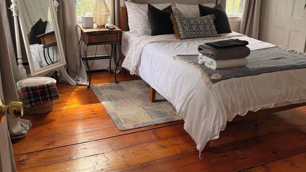 A bedroom with a bed, a bedside table, a mirror, a window with curtains, and a rug on the wooden floor. - Mariam's On Becket (Salem)
