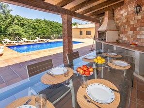 An outdoor dining area with a glass table, chairs, and a view of a swimming pool and greenery.
