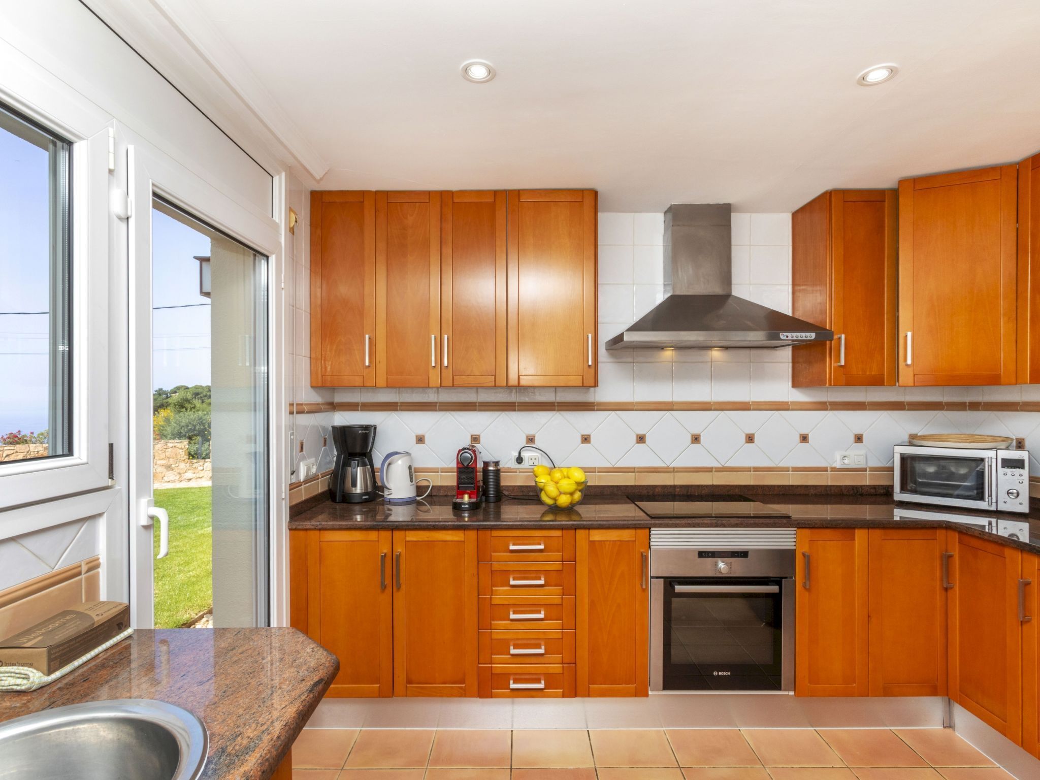 A modern kitchen with wooden cabinets, a stainless steel range hood, a Bosch oven, and a window providing a view of the outdoors.