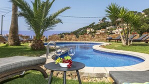 Ein Poolbereich mit Liegestühlen, einem Tisch mit Obstschale und Blick auf das Meer und die Hügel im Hintergrund.
