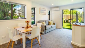 Una sala de estar moderna con una mesa de comedor, sillas blancas, un sofá gris con cojines y un ventanal con vistas al exterior.