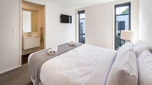 A hotel room with a bed, a TV mounted on the wall, a bathroom with a mirror and towel, and a window with a view of the outside.
