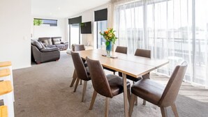 A modern living room with a sofa, a dining table with chairs, and a large window with sheer curtains.
