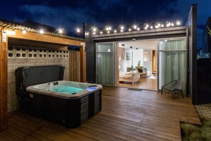 A hot tub on a wooden deck with a covered patio area and a sliding glass door leading to a living room.