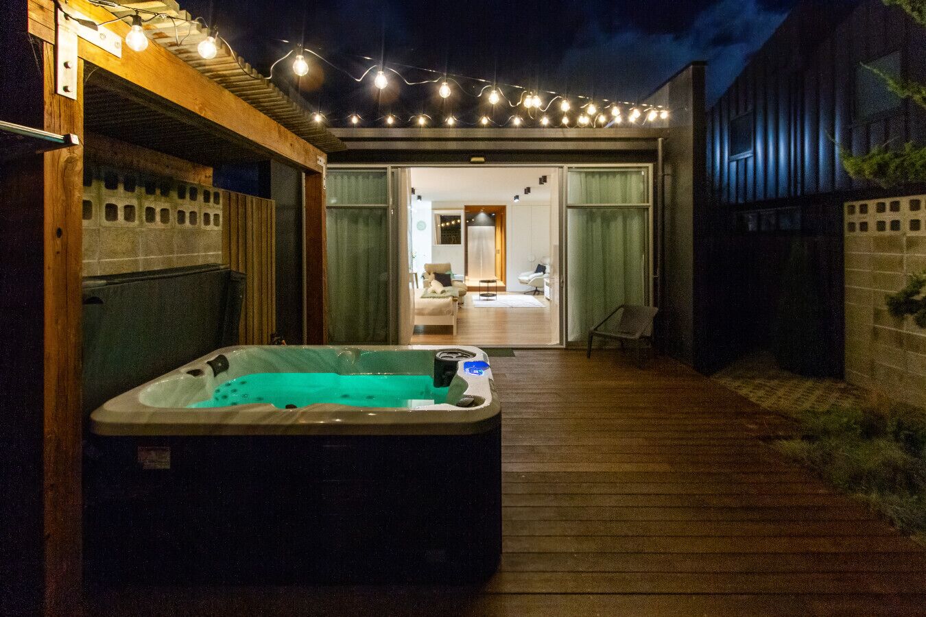 Ein Whirlpool in einem überdachten Außenbereich mit Holzterrasse, verbunden mit einem Gebäude mit einer Glaswand, die in ein Wohnzimmer führt.