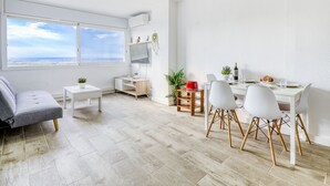 Ett modernt och väl upplyst vardagsrum med ett stort fönster, en grå soffa, en matplats med vita stolar och ett bord samt ett litet köksområde med en röd köksapparat.
