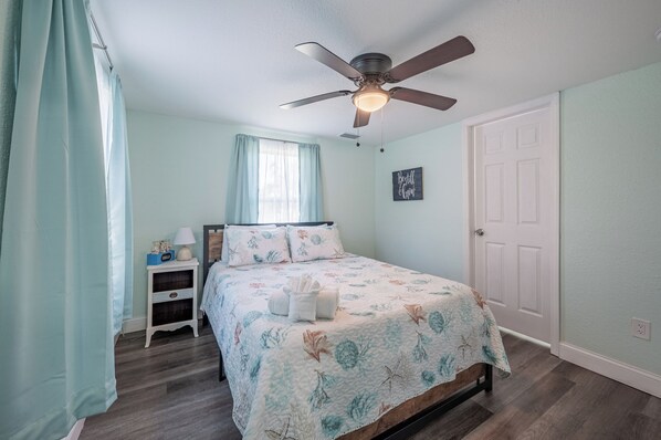 Un dormitorio con cama, mesita de noche, ventilador de techo, ventana con cortinas y puerta.