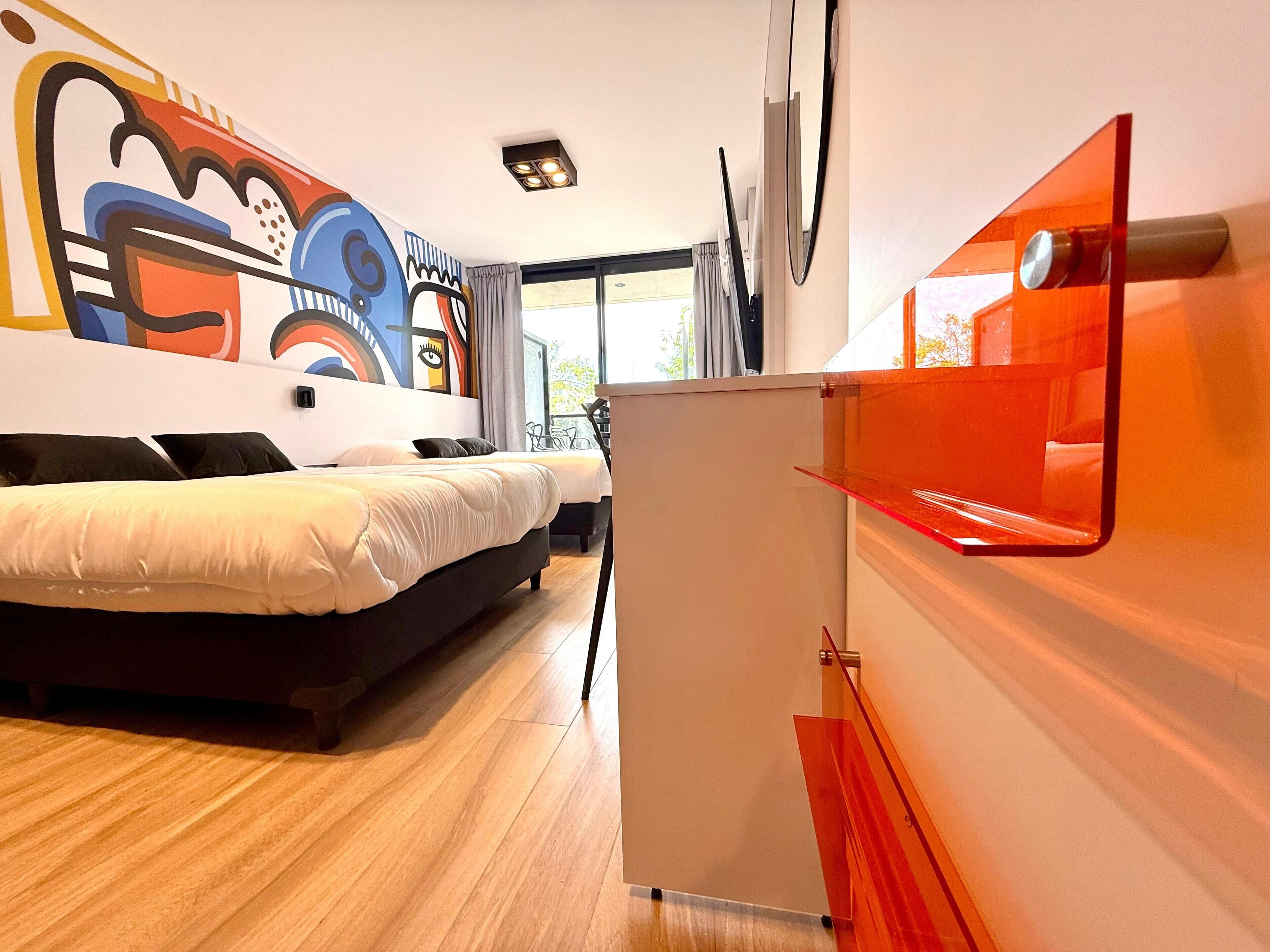 A modern hotel room with a large bed, a small desk, a colorful abstract mural, and a view of the outdoors.