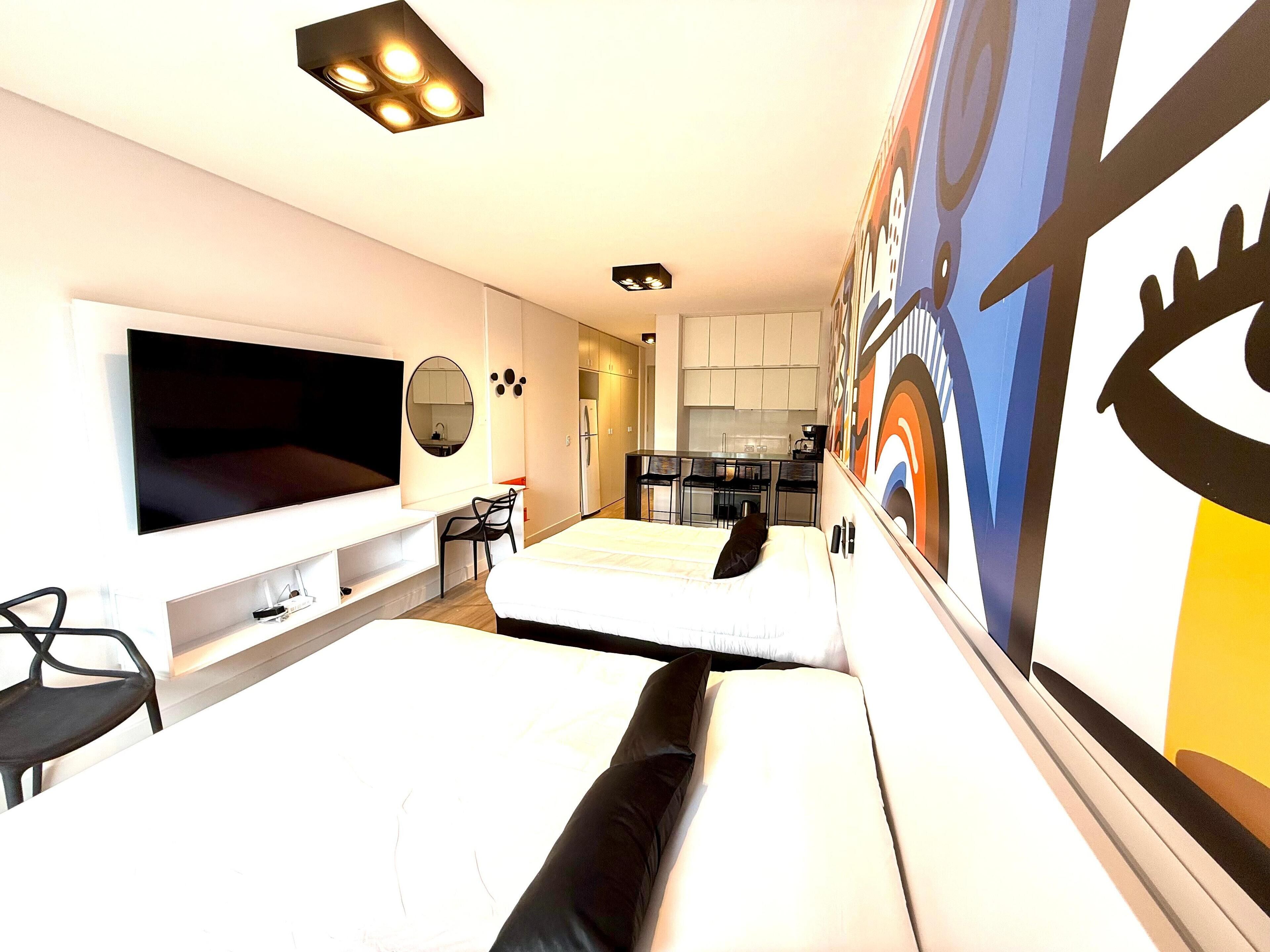 A modern hotel room with a large flat-screen TV, a desk with a chair, a bed with black bedding, and a kitchen area visible through an open door.