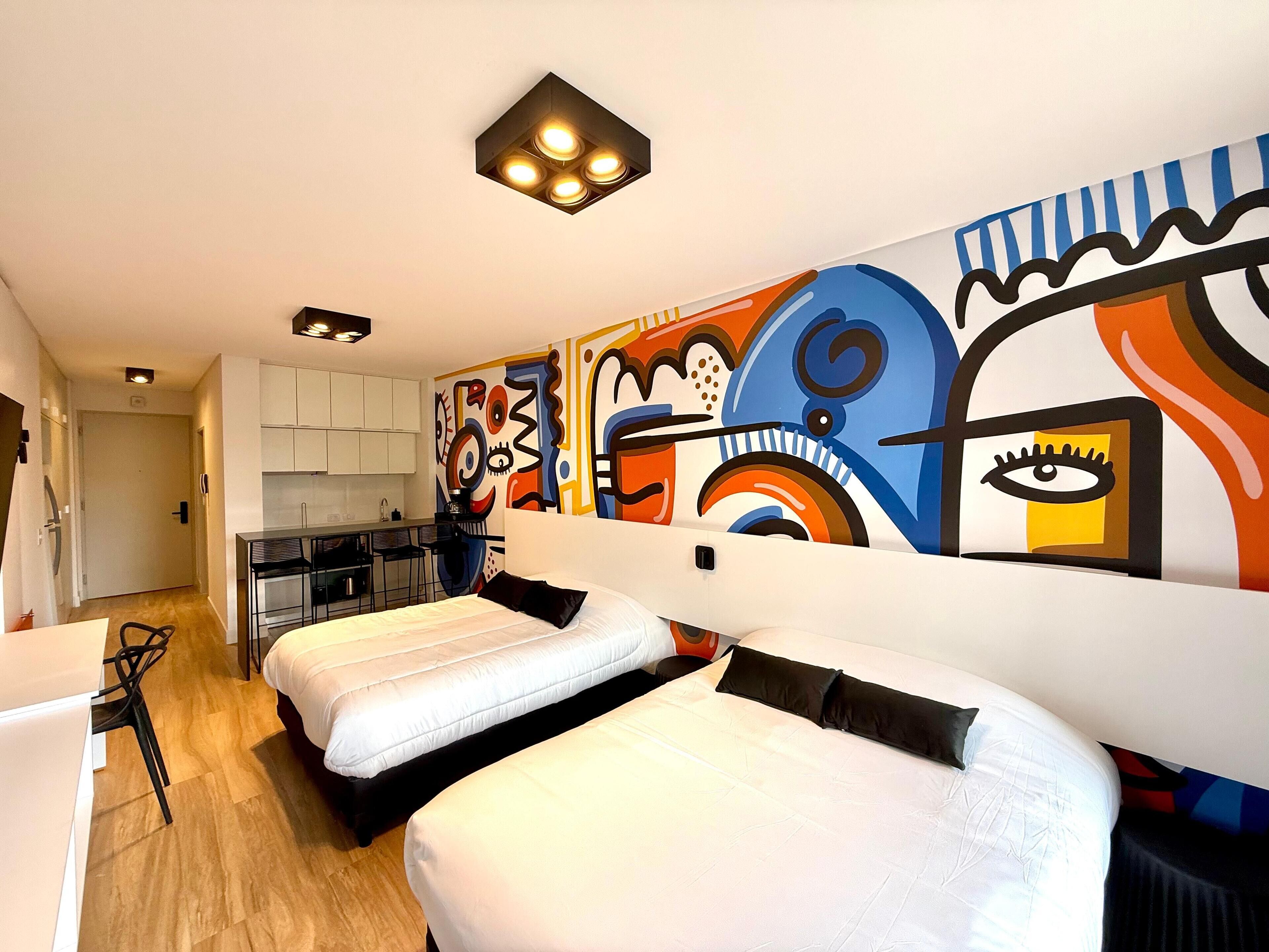 A modern hotel room with a kitchen area, two beds, and a colorful abstract mural on the wall.
