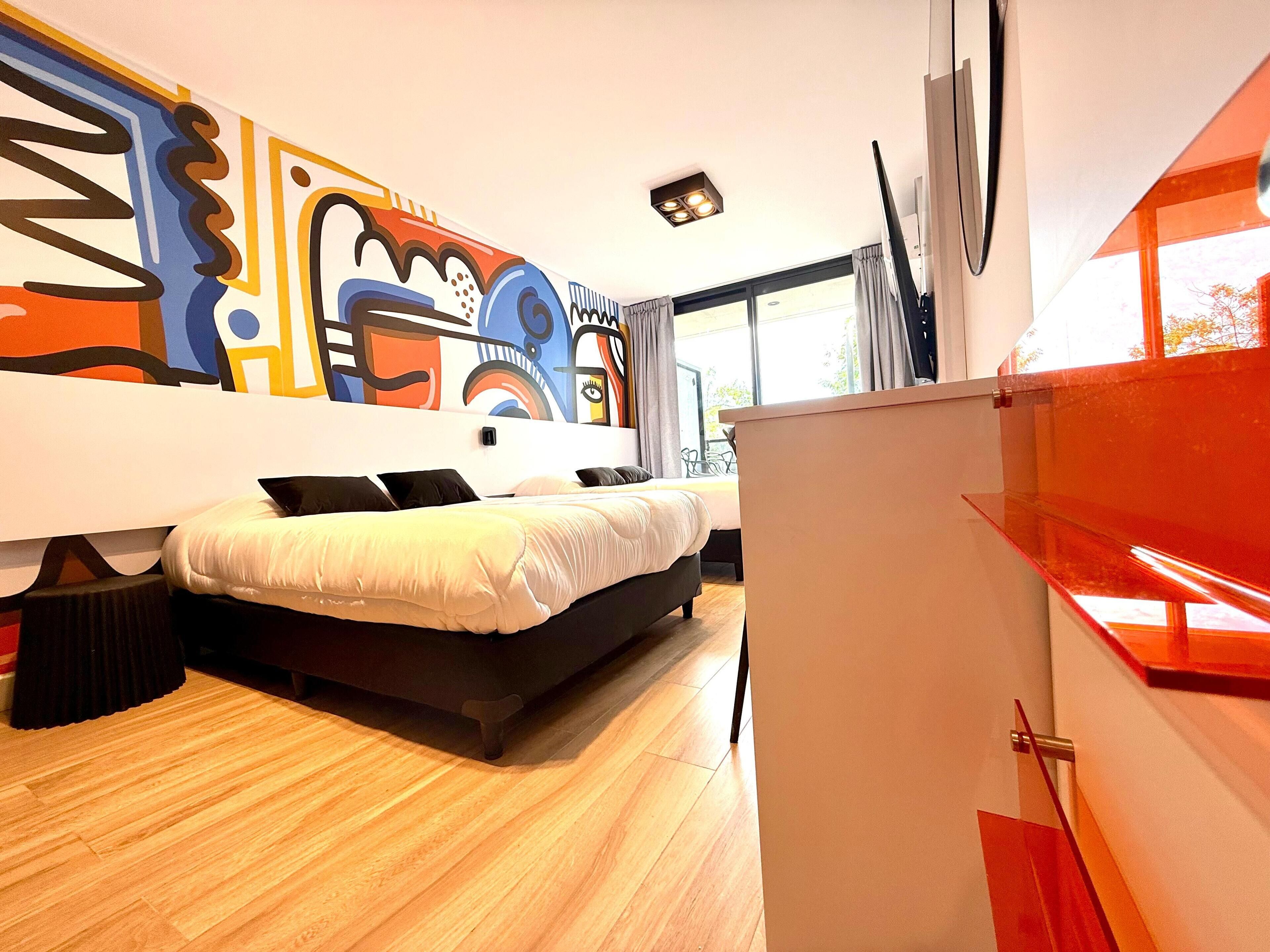 Een moderne slaapkamer met een groot bed, een bureau en een kleurrijke abstracte muurschildering.