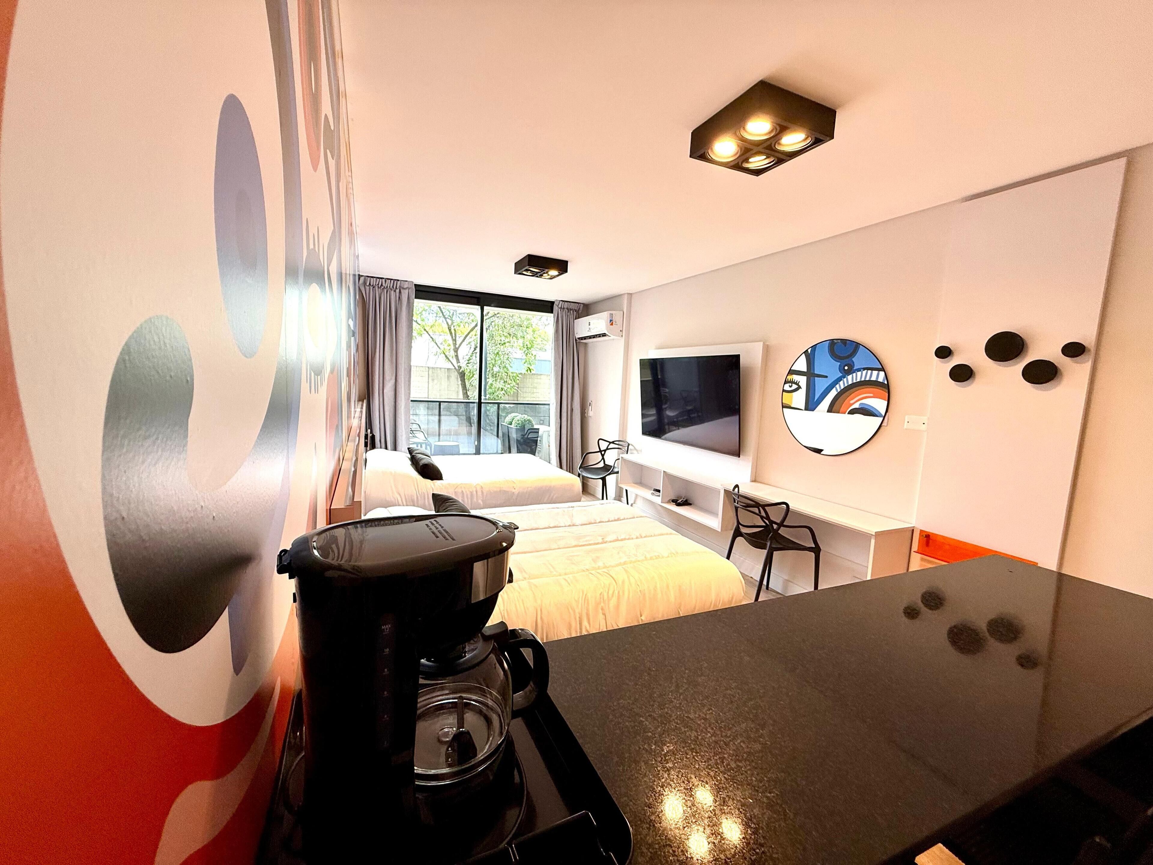 Una habitación de hotel moderna con cama, cafetera, televisor y un ventanal grande.