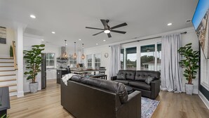 A spacious living room with a dark brown leather sofa, a ceiling fan, and large windows with curtains.
