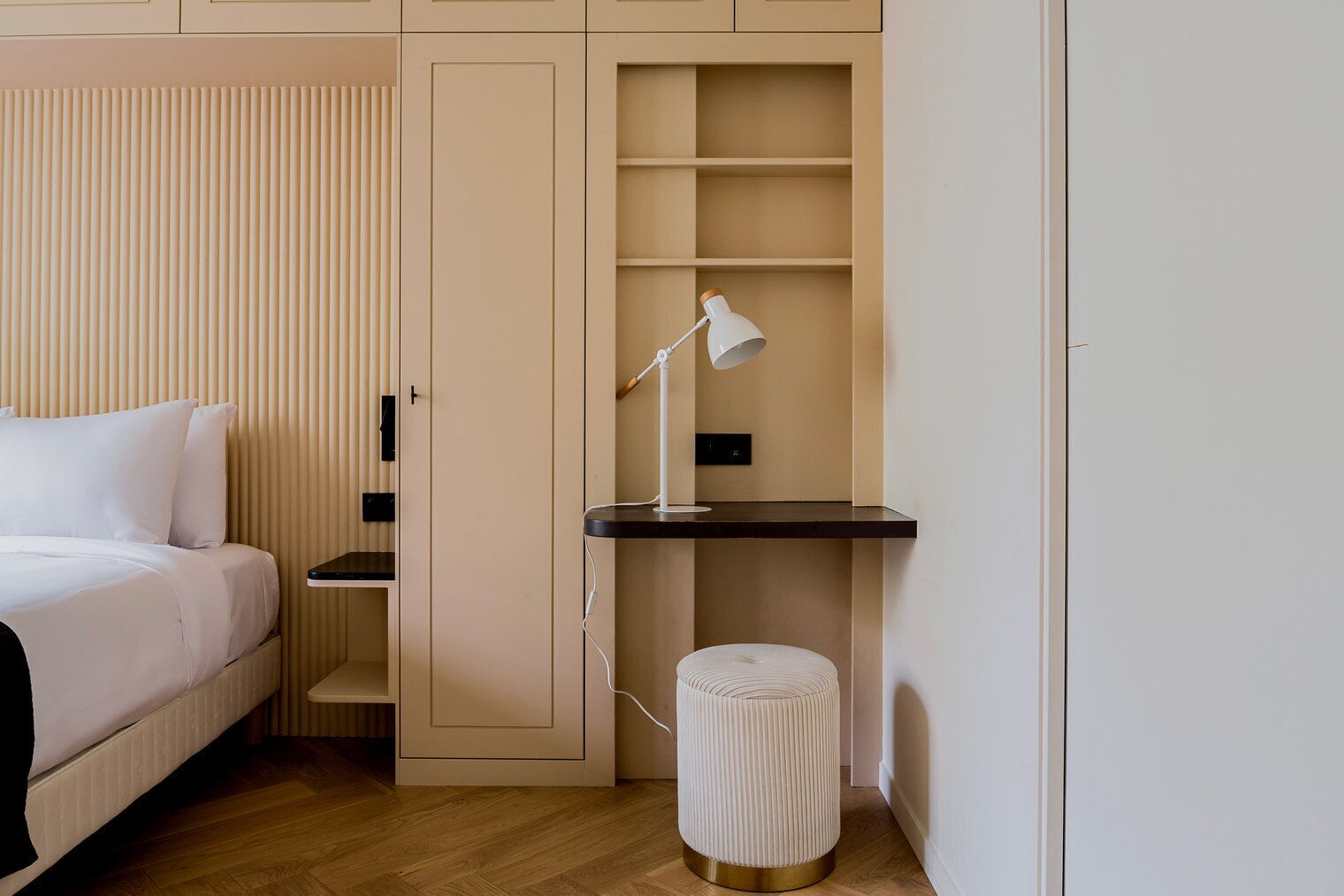 A bedroom with a bed, a desk with a lamp, a small stool, and built-in shelving.