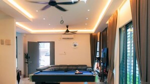 Een moderne woonkamer met een pooltafel, plafondventilator en grote ramen voorzien van gordijnen.