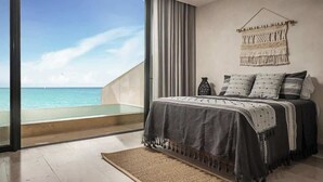 Een slaapkamer met een groot raam dat uitkijkt op de oceaan, een macramé wanddecoratie en een bed met kussens met een patroon.
