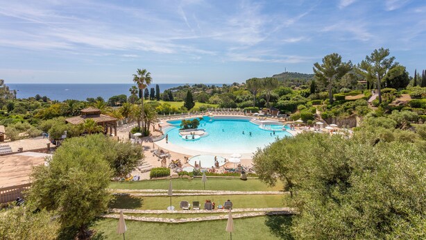 A resort with a large swimming pool, lush greenery, and a view of the sea.