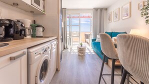 A modern kitchen with a washing machine, a dining area with a blue sofa and chairs, and a view through a glass door.