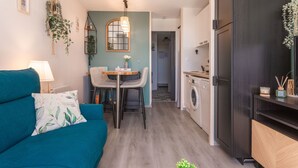 A compact living space with a sofa, dining table, chairs, a hanging plant, a washing machine, and a kitchen area.