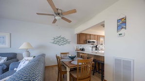 A modern living room with a sofa, dining table, chairs, a ceiling fan, kitchen area with cabinets and appliances, and a wall-mounted artwork. - 4203 - Here Comes the Sun - Duck Oceanside Condo | Community Pool & Beach (Duck)