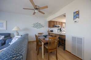 Uma sala de estar moderna com sofá, mesa de jantar, cadeiras, ventilador de teto, área de cozinha com armários e eletrodomésticos, e uma obra de arte pendurada na parede.