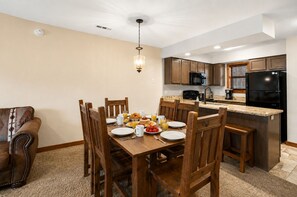 A dining area with a wooden table set for breakfast, a kitchen with modern appliances, and a ceiling light fixture.