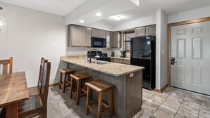 A modern kitchen with a granite countertop, wooden stools, a stainless steel refrigerator, and a microwave. - Table Rock Lakefront  Condo • Sleeps 10 • Balcony • 2 Min to Silver Dollar City (Branson)