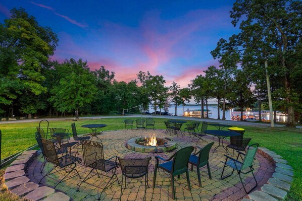 A circular outdoor seating area with a fire pit, surrounded by chairs and tables, overlooking a body of water.