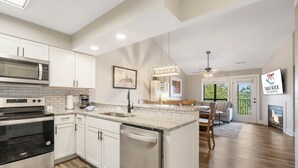 A modern kitchen with white cabinets, a built-in microwave, and a dishwasher. There is a dining area with a table and chairs, and a fireplace. - Walk to Table Rock Lake • Family Friendly • Minutes to SDC (Branson)