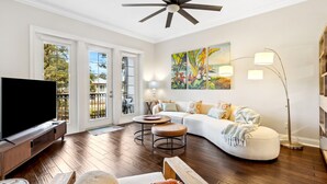 Ein Wohnzimmer mit Flachbildfernseher, Holzboden, Deckenventilator, großem Fenster, einer Couch und einer Stehlampe.