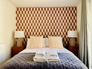 Habitación de hotel con cabecera estampada, dos mesitas de noche con lámparas, cama con cubrecama gris y dos toallas dobladas sobre la cama.