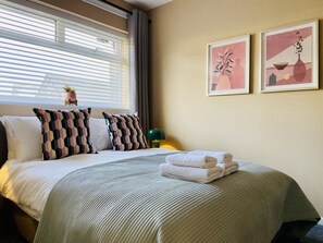 Ein ordentlich bezogenes Bett mit einer gestreiften Tagesdecke, zwei Kissen und gefalteten Handtüchern an der Kopfteil. An der Wand hängen zwei gerahmte Kunstwerke, ein Fenster mit Jalousien und ein kleiner Topfpflanze auf dem Fensterbrett.