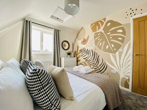Ein Schlafzimmer mit einem Bett, Kissen, einem Nachttisch, einem Fenster mit Vorhängen, einer Wand mit Blättermotiv und einer Tür.