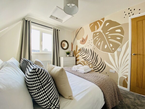 Un dormitorio con cama, almohadas, una mesita de noche, una ventana con cortinas, una pared con diseño de hojas y una puerta.
