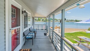 A covered porch with a view of a lake and beach, featuring a bench, a table, and a railing.