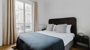 Une chambre à coucher avec un grand lit, une table de chevet, une lampe et une fenêtre avec des rideaux.