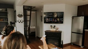 A modern living room with a fireplace, a television displaying a movie, and a kitchen area with a refrigerator.