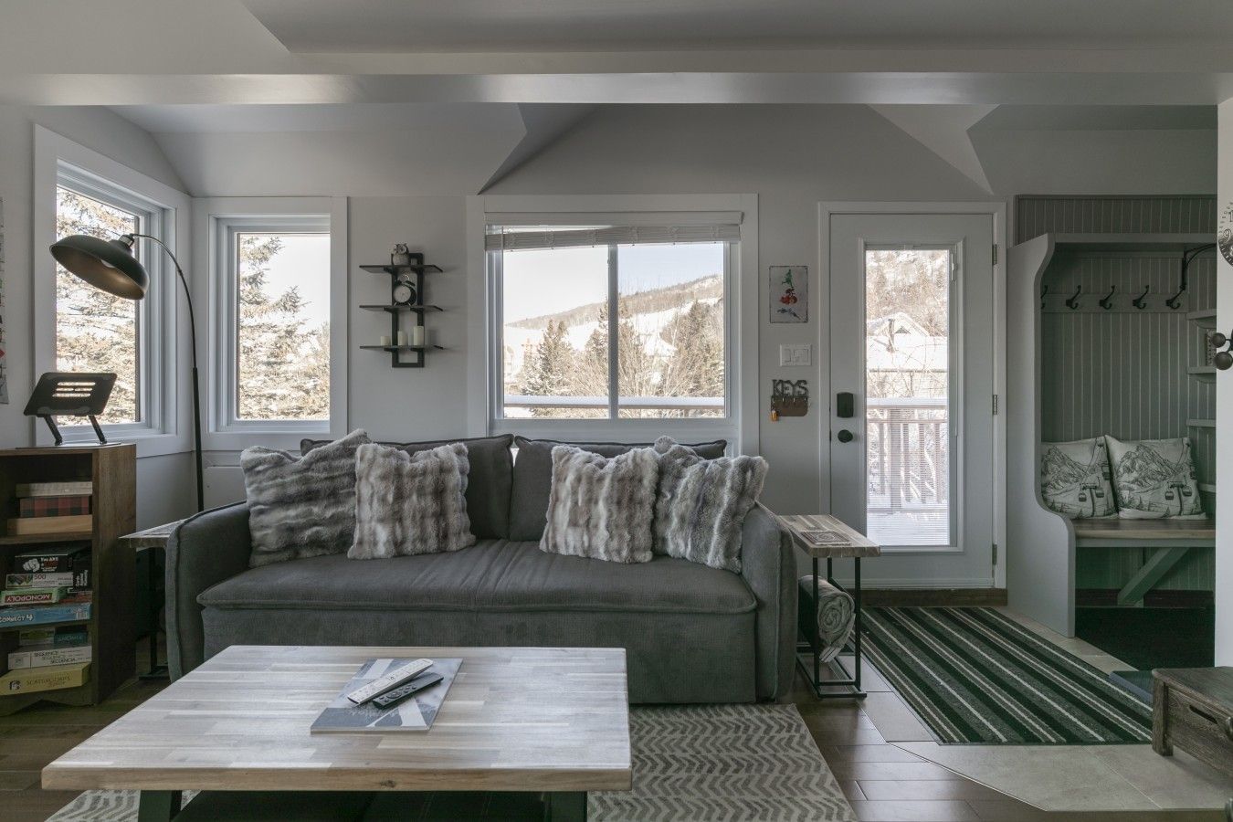 A living room with a grey sofa, a coffee table, and a bookshelf. There are two windows with blinds, a door, and a coat rack.