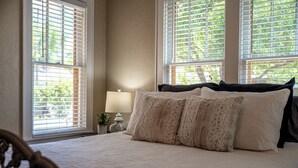 Une chambre à coucher avec un lit, deux fenêtres équipées de stores, une table de chevet avec une lampe et une petite plante.