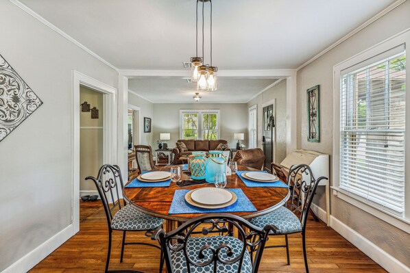 A dining room with a wooden table, chairs, and a chandelier. There is a living room in the background with a couch and a coffee table. - Cornerstone Cottage-Near Downtown-Pet Friendly (Stephenville)