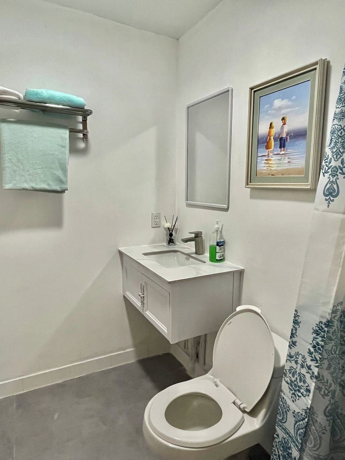 Een badkamer met toilet, wastafel en spiegel. Er is een handdoekhouder met een handdoek en een foto in een lijst aan de muur.