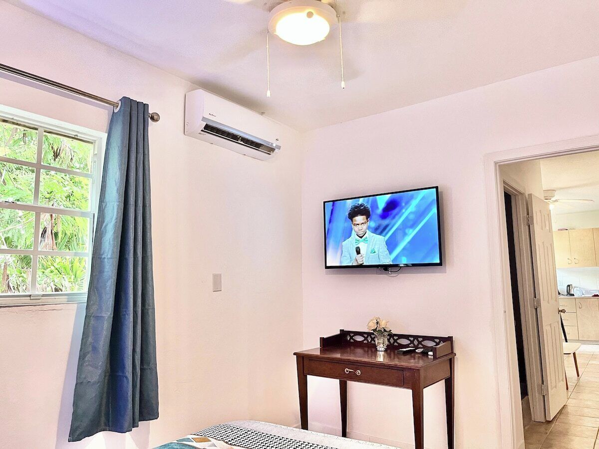 Een kamer met een bed, een televisie aan de muur, een kleine tafel met een vaas en een raam met gordijnen.