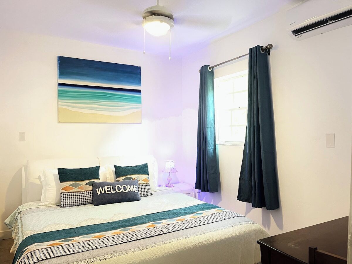 A bedroom with a bed, a welcome pillow, a painting, a window with curtains, and a ceiling fan.
