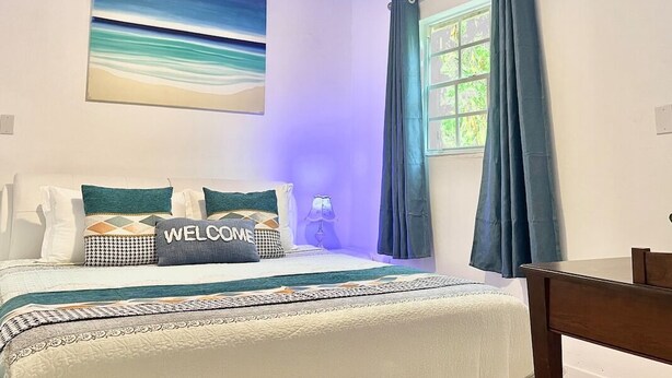 Une chambre à coucher avec un lit, une table de chevet, une lampe, une fenêtre avec des rideaux et un tableau au mur.