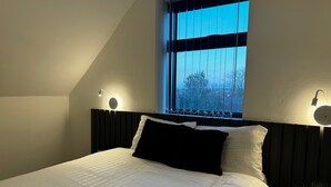 Ett sovrum med en säng, två sänglampor, ett fönster med utsikt över träd och en växt på nattduksbordet.