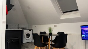 Ett kompakt kök med tvättmaskin, en matplats med bord och stolar, en liten papperskorg och en tv som är monterad på väggen.