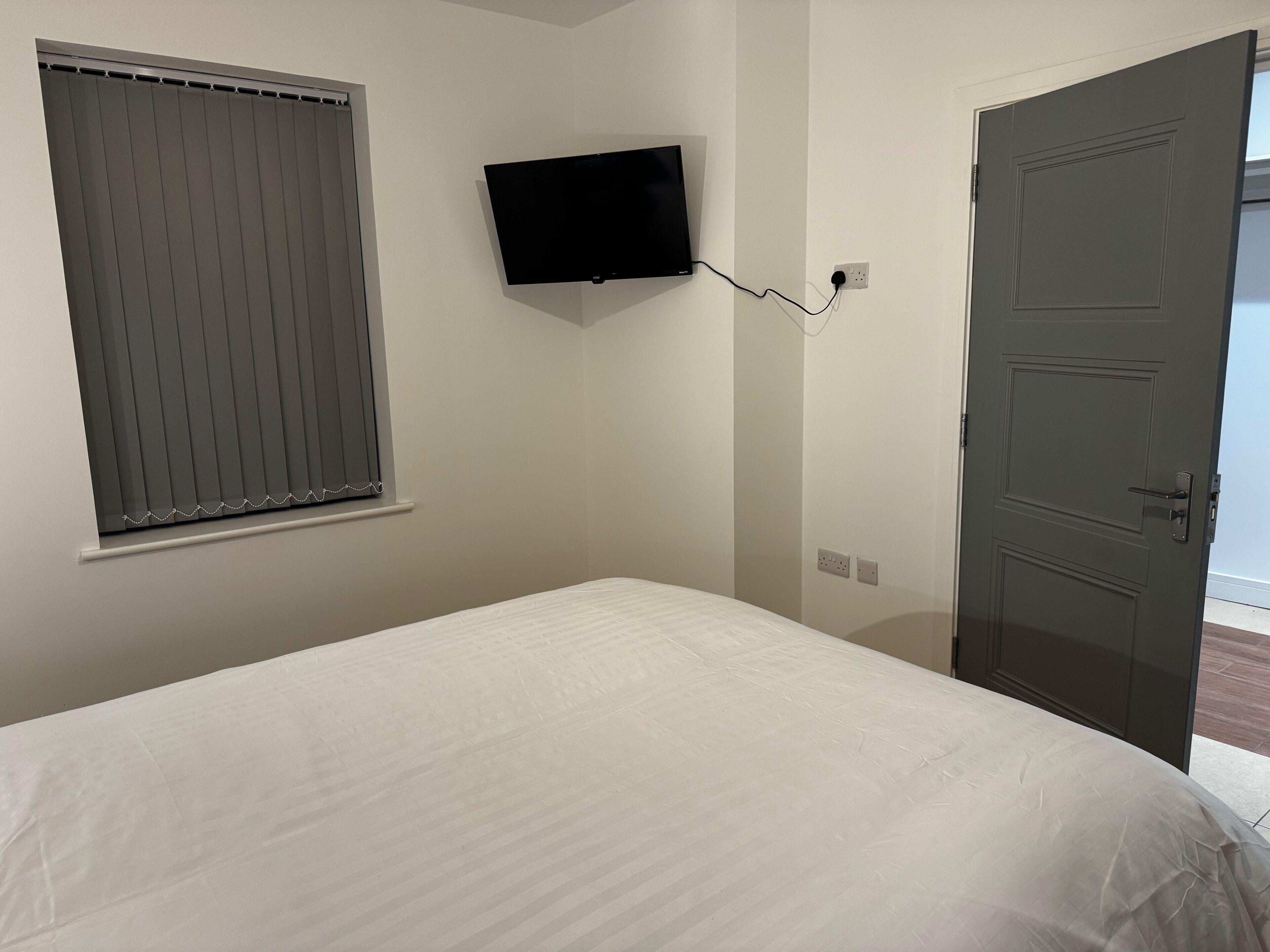 Un dormitorio con cama, televisor montado en la pared, ventana con persianas y una puerta.