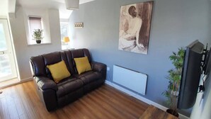 En stue med en mørk lædersofa, trægulv, et stort abstrakt maleri, en radiator og et fjernsyn.