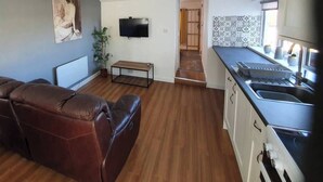 En kompakt opholdsstue med sofa, fjernsyn, et lille bord og et tekøkken med vask og bordplade.