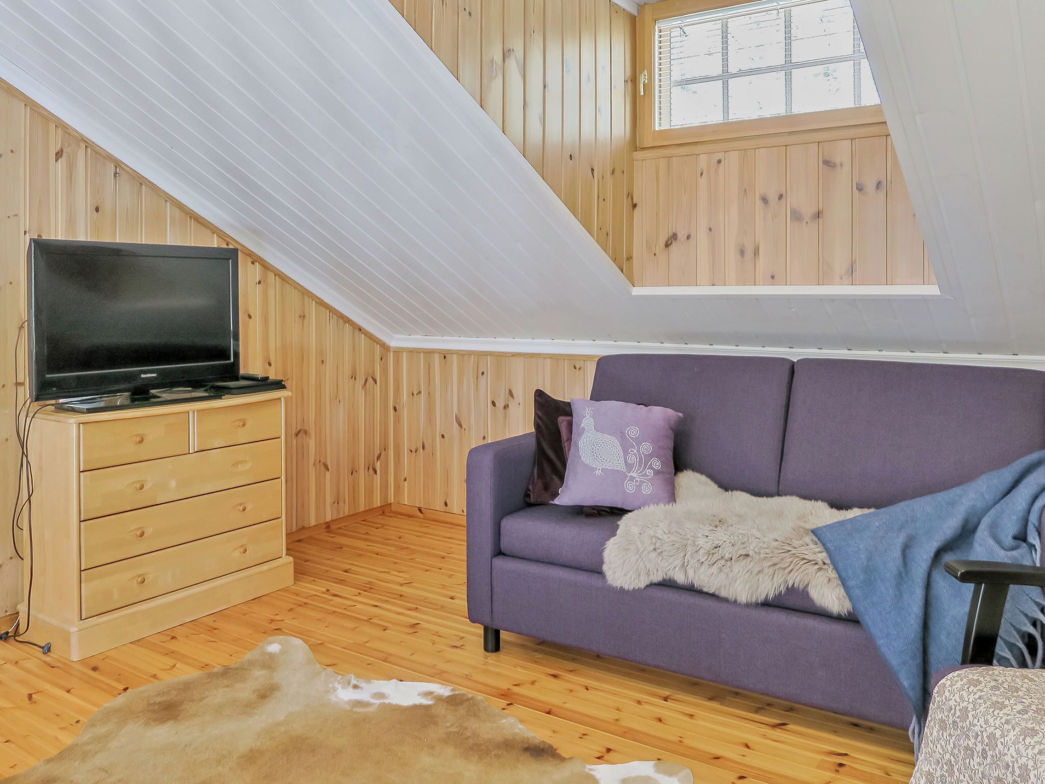 A loft-style room with a wooden floor, a purple sofa, a television on a wooden cabinet, and a large window.